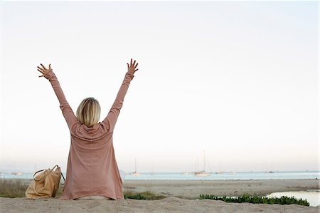 Blond woman sitting on a sandy beach, arms raised. Photographie de stock - Premium Libres de Droits, Code: 6118-08313701