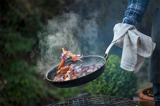 An outdoor cookout. A man holding a frying pan sauteeing vegetables above an open fire. Foto de stock - Sin royalties Premium, Código de la imagen: 6118-08521663