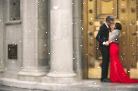 A woman in a long red evening dress with fishtail skirt and a fur stole, and a man in a suit kissing on the steps of a building. Stock Photo - Premium Royalty-Free, Code: 6118-08521887