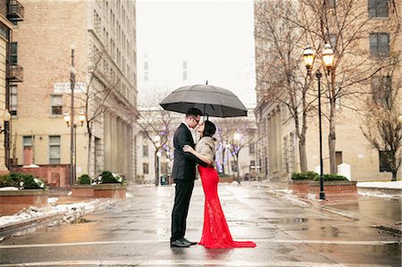 A woman in a long red evening dress with fishtail skirt and a fur stole, and a man in a suit, kissing under an umbrella in the city. Stock Photo - Premium Royalty-Free, Code: 6118-08521838