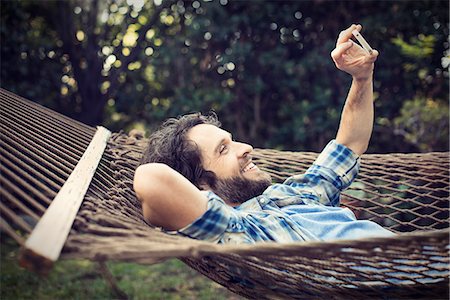 A man lying in a garden hammock taking selfies with his phone. Stock Photo - Premium Royalty-Free, Code: 6118-08521955