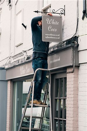 simsearch:6118-08659688,k - A man on a ladder fixing a painted name sign onto a bracket on a shopfront. Stockbilder - Premium RF Lizenzfrei, Bildnummer: 6118-08659693