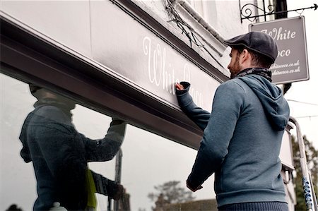 road working - A man on a ladder fixing a painted name sign onto a bracket on a shopfront. Stock Photo - Premium Royalty-Free, Code: 6118-08659695