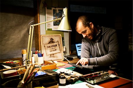 A man in his workshop using a digital tablet with a touch screen surrounded by equipment for signwriting and screen-printing. Fotografie stock - Premium Royalty-Free, Codice: 6118-08659700