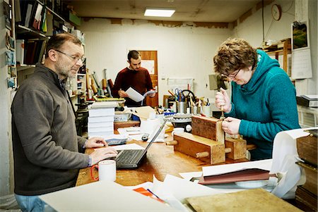 A woman working on the spine of a bound book with a hand tool. A man using a laptop computer. Stock Photo - Premium Royalty-Free, Code: 6118-08659718