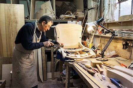 simsearch:6118-08660112,k - Man standing at a work bench in a carpentry workshop, working on a wooden chair with a small hand saw. Foto de stock - Royalty Free Premium, Número: 6118-08660097