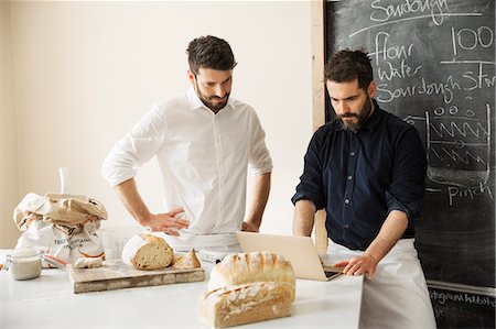fait maison - Two bakers standing at a table, using a laptop computer, freshly baked bread, a blackboard on the wall. Photographie de stock - Premium Libres de Droits, Code: 6118-08660015