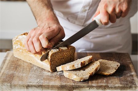 simsearch:6118-09079256,k - Close up of a baker slicing a freshly baked loaf of bread with a bread knife. Stock Photo - Premium Royalty-Free, Code: 6118-08660030