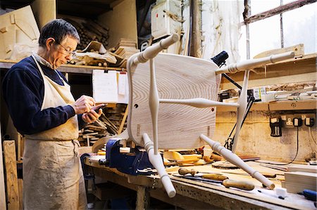 Man standing at a work bench in a carpentry workshop, working on a wooden chair. Stock Photo - Premium Royalty-Free, Code: 6118-08660102