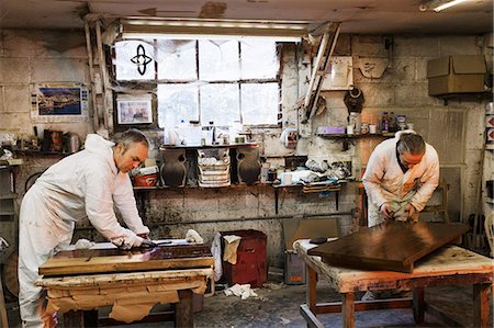 Man standing in a carpentry workshop, wearing protective suit and gloves, applying varnish onto a wooden surface with a cloth. Fotografie stock - Premium Royalty-Free, Codice: 6118-08660108