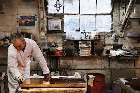 staining wood brush - Man standing in a carpentry workshop, wearing protective suit and gloves, applying varnish onto a wooden surface with a cloth. Stock Photo - Premium Royalty-Free, Code: 6118-08660109