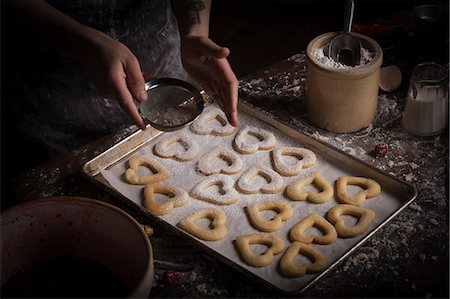 simsearch:6118-08660146,k - Valentine's Day baking, woman sprinkling icing sugar over heart shaped biscuits on a baking tray. Stock Photo - Premium Royalty-Free, Code: 6118-08660127