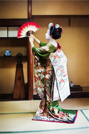 A woman dressed in the traditional geisha style, wearing a kimono and obi, with an elaborate hairstyle and floral hair clips, with white face makeup with bright red lips and dark eyes. Standing in a classic pose with fan raised, side view. Stock Photo - Premium Royalty-Free, Code: 6118-08761742