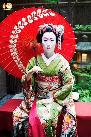 A woman dressed in the traditional geisha style, wearing a kimono and obi, with an elaborate hairstyle and floral hair clips, with white face makeup with bright red lips and dark eyes seated holding an umbrella. Stock Photo - Premium Royalty-Free, Code: 6118-08761746