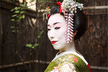 A woman dressed in the traditional geisha style, wearing a kimono and obi, with an elaborate hairstyle and floral hair clips, with white face makeup with bright red lips and dark eyes. Stock Photo - Premium Royalty-Free, Code: 6118-08761749