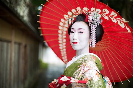 A woman dressed in the traditional geisha style, wearing a kimono and obi, with an elaborate hairstyle and floral hair clips, with white face makeup with bright red lips and dark eyes holding a red paper parasol. Stock Photo - Premium Royalty-Free, Code: 6118-08761758