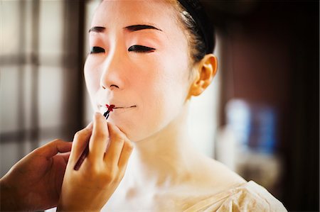 A modern woman creating the traditional geisha vivid red lips by painting on lipstick with a fine brush. White face makeup. Stock Photo - Premium Royalty-Free, Code: 6118-08761789