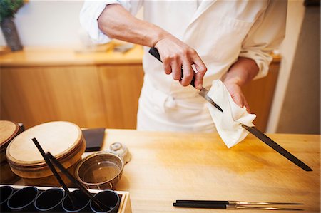 simsearch:6118-08762017,k - A chef working in a small commercial kitchen, an itamae or master chef preparing to make sushi, cleaning his knife. Photographie de stock - Premium Libres de Droits, Code: 6118-08761874