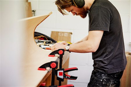 A furniture workshop making bespoke contemporary furniture pieces using traditional skills in modern design. A  man at a workbench working on a piece of curved wood held with clamps. Stock Photo - Premium Royalty-Free, Code: 6118-08725442