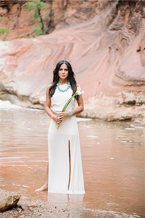 Young woman with long brown hair, wearing a long white dress, standing by a river, holding Arum Lilies. Photographie de stock - Premium Libres de Droits, Code: 6118-08725559