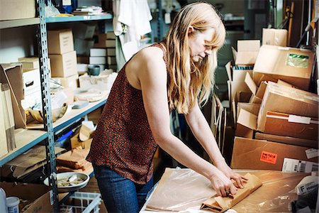 Woman with long blond hair standing in the store room of a shop, wrapping merchandise in brown paper. Stock Photo - Premium Royalty-Free, Code: 6118-08725762