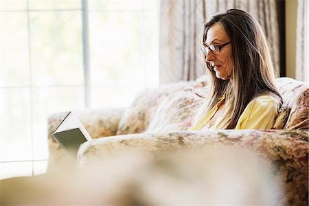 Senior woman with long brown hair sitting on a sofa, reading a book, wearing reading glasses. Stock Photo - Premium Royalty-Free, Code: 6118-08725765
