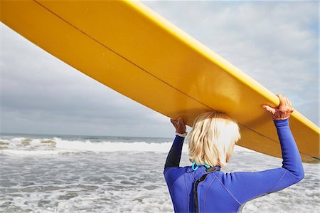 senior woman alone from behind - Senior woman on a beach, wearing a wetsuit and carrying a surfboard on her head. Stock Photo - Premium Royalty-Free, Code: 6118-08726073