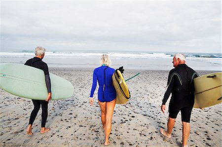 simsearch:6118-08129644,k - Senior woman and two senior men on a beach, wearing wetsuits and carrying surfboards. Fotografie stock - Premium Royalty-Free, Codice: 6118-08726072