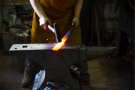 A blacksmith strikes a length of red hot metal on an anvil with a hammer in a workshop. Photographie de stock - Premium Libres de Droits, Code: 6118-08729044