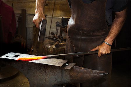 A blacksmith strikes a length of red hot metal on an anvil with a hammer in a workshop. Stock Photo - Premium Royalty-Free, Code: 6118-08729033