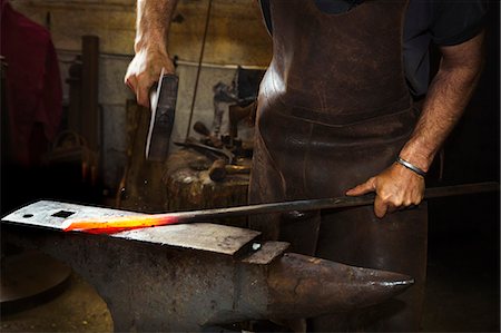 A blacksmith strikes a length of red hot metal on an anvil with a hammer in a workshop. Photographie de stock - Premium Libres de Droits, Code: 6118-08729033