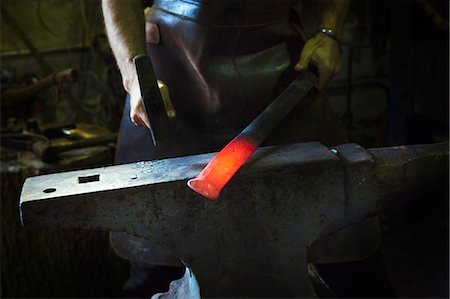 A blacksmith strikes a length of red hot metal on an anvil with a hammer in a workshop. Stock Photo - Premium Royalty-Free, Code: 6118-08729034