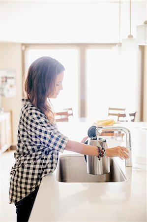 simsearch:6118-08729154,k - Woman with long brown hair, wearing a chequered shirt, standing at a kitchen sink. Foto de stock - Sin royalties Premium, Código: 6118-08729145