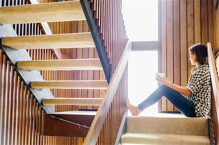 Woman with long brown hair sitting on a wooden staircase, looking out of window. Stock Photo - Premium Royalty-Free, Code: 6118-08729166