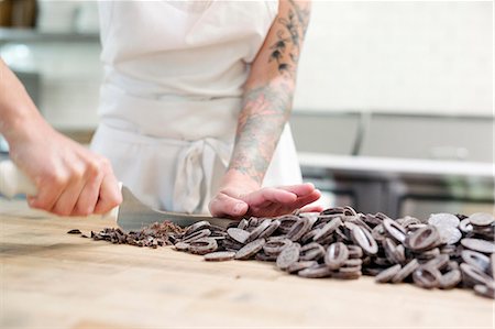 Close up of a woman wearing a white apron standing at a work counter in a bakery, chopping chocolate. Stock Photo - Premium Royalty-Free, Code: 6118-08729118