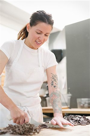 Woman wearing a white apron standing at a work counter in a bakery, chopping chocolate. Stock Photo - Premium Royalty-Free, Code: 6118-08729120