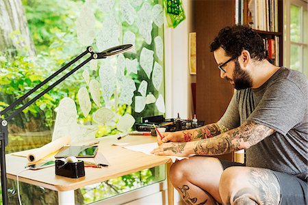 Bearded man with tattoos on his arms and legs sitting at a desk, drawing. Fotografie stock - Premium Royalty-Free, Codice: 6118-08729331