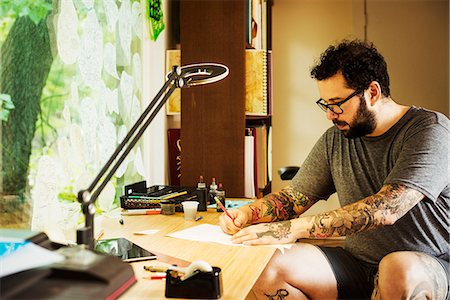 Bearded man with tattoos wearing glasses, sitting at a desk, drawing. Fotografie stock - Premium Royalty-Free, Codice: 6118-08729332