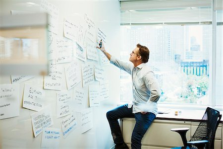 simsearch:6118-09079486,k - A man leaning on a desk in an office looking at pieces of paper pinned on a whiteboard. Foto de stock - Royalty Free Premium, Número: 6118-08842124