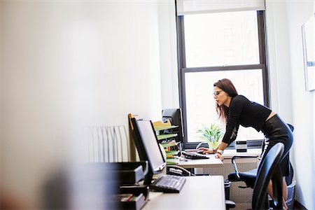 A young woman standing at a desk looking at a computer screen. Stock Photo - Premium Royalty-Free, Code: 6118-08842207