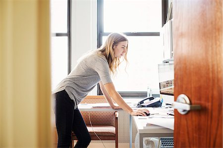 A young woman in an office standing over a desk and working at a computer. Stock Photo - Premium Royalty-Free, Code: 6118-08842210