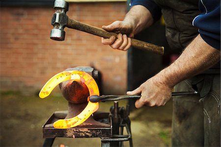 A farrier using tongs and hammer to hold and shape a red glowing heated metal horseshoe to be fitted. Stockbilder - Premium RF Lizenzfrei, Bildnummer: 6118-08882963