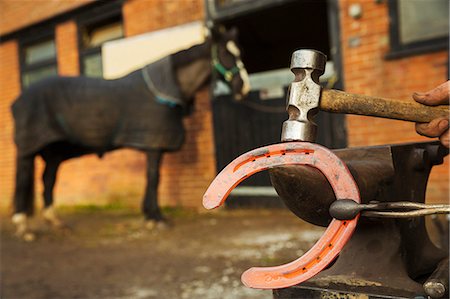 A farrier using tongs and hammer to hold and shape a red glowing heated metal horseshoe to be fitted. Photographie de stock - Premium Libres de Droits, Code: 6118-08882978