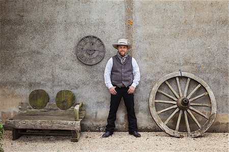 simsearch:6118-08947762,k - Bearded man wearing Stetson standing with his hands in his pockets outside a barn next to a wooden bench and large wooden carriage wheel. Stockbilder - Premium RF Lizenzfrei, Bildnummer: 6118-08947758