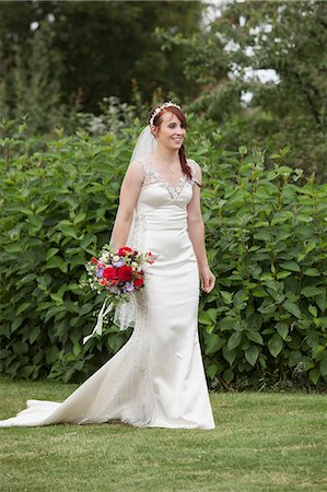 simsearch:6118-07521749,k - Bride in her wedding dress standing in a garden, holding wedding bouquet of red flowers. Photographie de stock - Premium Libres de Droits, Code: 6118-08947760