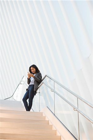 A woman standing on a stairway in the Oculus building at the World Trade Centre under a dramatic ridged white ceiling, checking her phone. Stock Photo - Premium Royalty-Free, Code: 6118-08971304