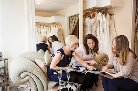 simsearch:6118-08971470,k - Three women in a wedding dress shop, one bride to be and two retail advisors looking through brochures and magazines. Two glasses of champagne on the table. Photographie de stock - Premium Libres de Droits, Code: 6118-08971448