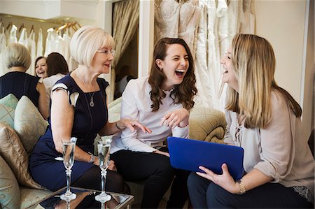 simsearch:6118-08971470,k - Three women in a wedding dress shop, one bride to be and two retail advisors looking at a digital tablet.  Two glasses of champagne on the table. Photographie de stock - Premium Libres de Droits, Code: 6118-08971449