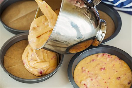 Close up high angle view of cake batter being poured from metal mixing bowl into round baking tins. Stock Photo - Premium Royalty-Free, Code: 6118-08971494