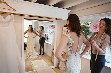 simsearch:6118-08971470,k - A young woman in a full length white wedding dress, looking at her reflection in the mirror in a bridal boutique. A dressmaker in the background. Photographie de stock - Premium Libres de Droits, Code: 6118-08971427